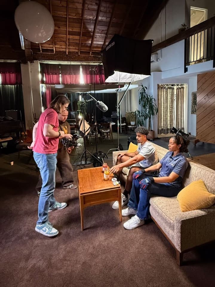 An interview in a loft studio: two people on a sofa, an overhead softbox and boom microphone, and the crew preparing the camera.