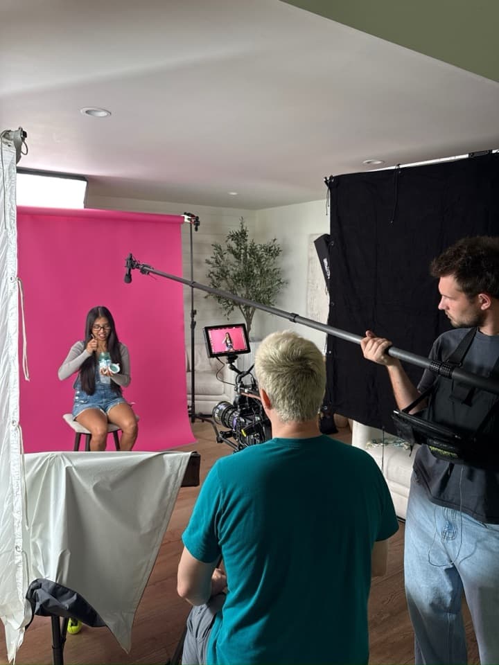 A presenter on a pink seamless backdrop, filmed on a cinema camera with a monitor, a boom operator holding the microphone overhead.
